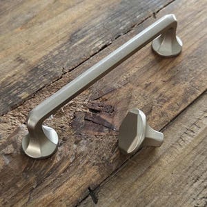 May include: A pair of brushed brass cabinet fittings. One is a long, straight handle with curved ends, and the other is a matching knob. The hardware is shown on a rustic wooden surface.