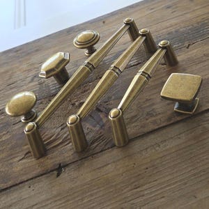 May include: A collection of antique brass cabinet hardware including three elongated drawer pulls with tapered centers and three distinct knobs featuring round, hexagonal, and square shapes with a brushed metallic finish.