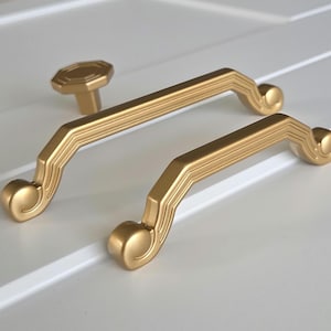 May include: Two gold-toned cabinet pulls with a decorative, ornate design. The pulls have a rounded, scroll-like shape at each end. A smaller, matching knob is also included.