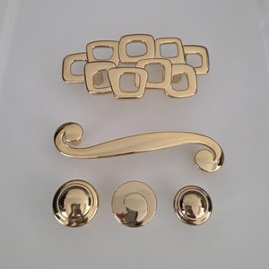 May include: A collection of gold-toned cabinet hardware, including a cluster of square-shaped pulls, a curved handle, and three round knobs. The hardware has a shiny, reflective surface, suggesting a modern or vintage aesthetic.
