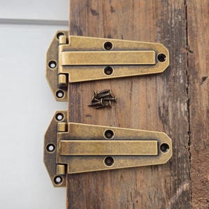 May include: Two antique brass-coloured hinges with screws, displayed on a weathered wooden surface. The hinges have a rectangular design with rounded edges and multiple screw holes. The screws are small and dark, contrasting with the lighter brass colour of the hinges.