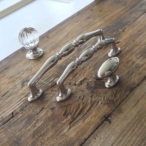 May include: A collection of cabinet hardware including two polished silver metal arched drawer pulls with a segmented design, one round clear glass knob with vertical ribbing, and one smooth oval-shaped polished silver metal cabinet knob.