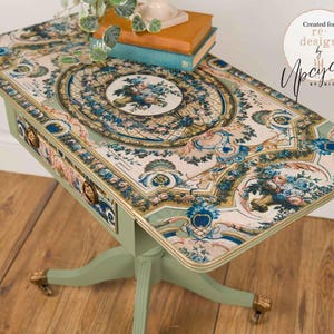 May include: A sage green pedestal table featuring a decorative floral transfer on the tabletop and drawer front. The design includes blue, gold, and cream patterns with a central floral medallion. It has two brass drawer pulls and claw feet.