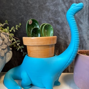 May include: A blue dinosaur-shaped planter with a wooden pot and green cactus-shaped spoons inside. The planter is sitting on a white surface.