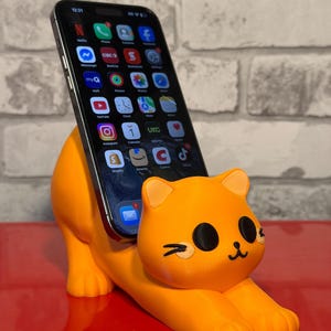 May include: Orange cat-shaped phone stand holding a black smartphone. The stand is made of a solid material and features a cute cat design with black eyes and a smiling mouth. The phone displays app icons.