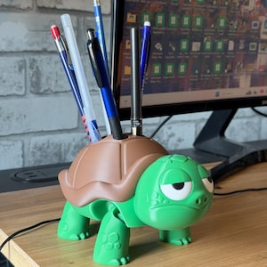 Op de afbeelding: Een groene en bruine schildpadvormige bureau-organizer met verschillende pennen en potloden. De schildpad heeft een chagrijnige uitdrukking en een bruin schild. De organizer is gemaakt van plastic en staat op een houten bureau.