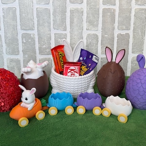 May include: A collection of Easter decorations and treats. Features a red decorative egg, a white basket filled with sweets, chocolate eggs with bunny ears, and a purple egg-shaped bunny. Also, a toy bunny in a carrot car and egg-shaped train carriages.