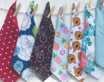 May include: Seven different fabric tea towels hanging on a clothesline. The towels are all different colors and patterns, including red with white polka dots, blue with white flowers, brown with bicycles, blue with pineapples, white with bees, blue with flowers, and blue with stripes.