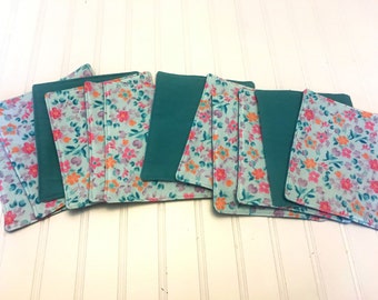 May include: A set of ten teal and floral fabric coasters. The coasters are made of two layers of fabric, one teal and one with a floral pattern in shades of pink, orange, and green.