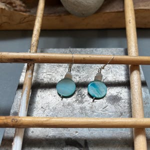 Puede incluir: Pendientes hechos a mano con discos redondos de concha azul-verde iridiscente. Cada pendiente tiene una pequeña cuenta de color claro y un gancho de alambre plateado. Los pendientes se exhiben sobre una superficie metálica con clavijas de madera.