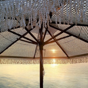 Puede incluir: Una sombrilla de playa de crochet blanco con estructura de madera, que proporciona sombra sobre una masa de agua con una puesta de sol dorada en el fondo.