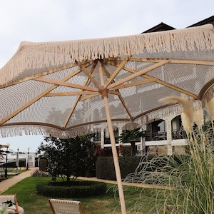 Könnte beinhalten: Ein großer, beigefarbener Strandschirm mit Fransen und einem Holzgestell. Der Schirm ist geöffnet und spendet Schatten über einem grasbewachsenen Bereich mit ein paar Palmen im Hintergrund.