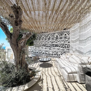 May include: A white sofa with striped cushions sits under a woven shade in a patio setting. A black and white patterned wall is behind the sofa. A small olive tree is in the foreground.