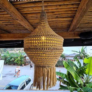 Op de afbeelding: Een grote, gehaakte, beige lampenkap hangt aan een houten plafond. De lampenkap heeft een franje en is van binnenuit verlicht. De lampenkap is gelabeld met "PATTERN No3".