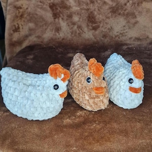 May include: Three handmade crochet chicken toys in white, tan, and light blue. Each chicken has orange accents for the comb and beak, and black button eyes. The chickens are made of a soft, textured yarn.