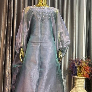 May include: A long, flowing, iridescent gray dress with a beaded neckline. The dress has a sheer, almost translucent fabric and is displayed on a mannequin against a backdrop of gold curtains.
