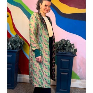 May include: A woman wearing a long, patterned coat with green and pink hues, paired with a white top and dark pants. The coat has a green cuff. The background features a colorful abstract mural and two dark blue planters.