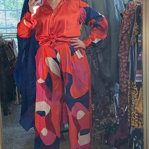 May include: A vibrant, two-piece outfit featuring a button-down shirt and wide-leg pants. The shirt is a solid orange, knotted at the waist. The pants have a bold pattern of red, pink, white, and navy blue abstract shapes. The outfit is made of a silky material.