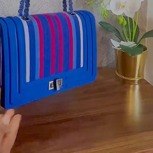 May include: A blue handbag with a flap closure and a silver clasp. The bag features vertical stripes in red, white, and blue. The chain strap is blue. A decorative plant in a gold pot sits on a wooden surface.