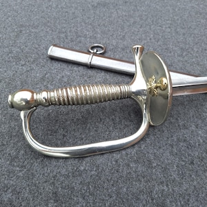 May include: A silver-toned sword with a ribbed handle, a curved hand guard, and a circular disc guard featuring a small gold-colored emblem. The sword is accompanied by a matching silver-toned metal scabbard with a single ring attachment.