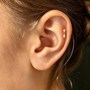May include: A gold-toned cartilage earring featuring a small round clear stone stud connected to a dangling marquise-shaped clear stone. The jewelry is positioned in the upper ear cartilage, showcasing a delicate metal chain link design.