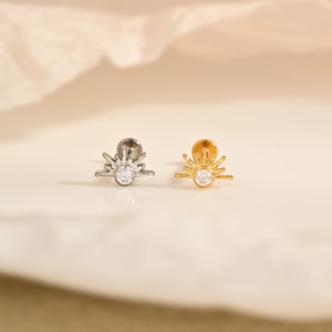 May include: Two stud earrings, one silver-toned and one gold-toned, each featuring a sunburst design. The center of each sunburst has a clear, round gemstone. The earrings are displayed on a white surface.