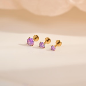 Puede incluir: Tres pendientes de botón con parte posterior plana, con piedras redondas de color púrpura iridiscente en engastes de cuatro garras. Los pendientes están dispuestos en fila, mostrando un poste de metal dorado pulido y un cierre plano circular.