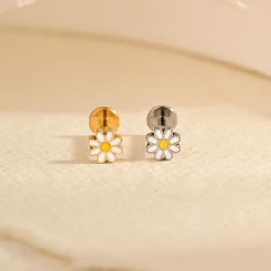 Peut inclure: Deux boucles d'oreilles à clous en forme de marguerite. L'une a une tige et un dos dorés, l'autre une tige et un dos argentés. Les deux présentent une marguerite en émail blanc avec un centre jaune.