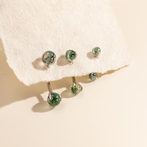Könnte beinhalten: Eine Sammlung silberfarbener Körperschmuckstücke mit kugelförmigen Moosachatsteinen mit grünen Einschlüssen. Das Set enthält zwei gebogene Barbell-Piercings mit Kugelenden und zwei passende Ohrstecker, die jeweils mit Metallkrallen gefasst sind.