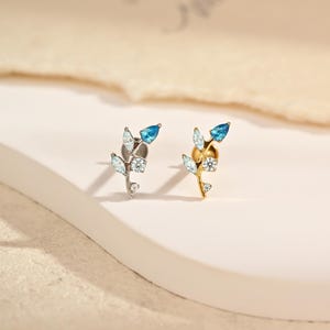 May include: Two stud earrings shaped like small branches, one in silver tone and one in gold tone. Each earring features clear marquise-cut stones, a round clear stone, and a single pear-cut blue stone at the tip, all set on a metal stem.