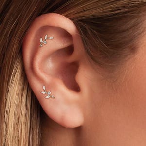 May include: Close-up of an ear adorned with two delicate, gold-toned leaf-shaped earrings. Each earring features small, sparkling gemstones, creating a subtle yet elegant design. The earrings are positioned on the upper and lower cartilage of the ear.