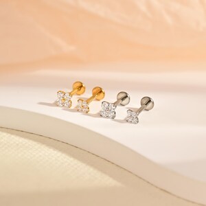 May include: Two pairs of stud earrings featuring a four-petal flower design. One pair is gold-toned and the other is silver-toned. Each flower is set with four small, clear, round-cut stones. The earrings have a flat back post and a round base.