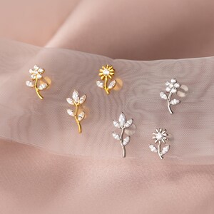 Könnte beinhalten: Eine Auswahl an Ohrsteckern mit floralen Motiven. Die Ohrringe zeigen Blumen- und Blattdesigns, verziert mit klaren Edelsteinen. Einige sind goldfarben, andere silberfarben, vor einem Hintergrund aus weichem rosa Stoff.