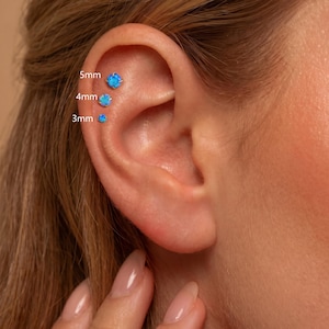 May include: Three round blue opal stud earrings in prong settings, arranged vertically along the upper ear cartilage. The studs are labeled with their respective sizes: 5mm, 4mm, and 3mm, indicating the diameter of each individual gemstone piece.