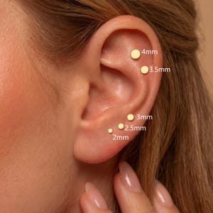 May include: Five gold-toned circular stud earrings arranged on an ear, labeled with sizes 2mm, 2.5mm, 3mm, 3.5mm, and 4mm. Each earring features a simple, flat, round design, showcasing a range of small diameters for multiple piercings.