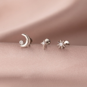 May include: Three silver-toned stud earrings. One earring is a crescent moon with a small star, another is a four-pointed star, and the third is a sunburst design. Each earring has a small, clear gemstone in the center.