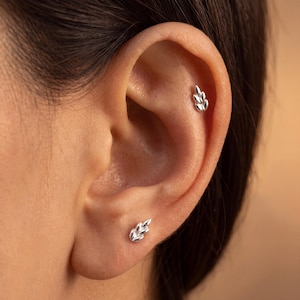 May include: A pair of silver-toned stud earrings, each featuring a small, three-leaf botanical design. One earring is positioned in the earlobe, while the other is placed in the upper cartilage area, both showcasing a polished metallic finish.