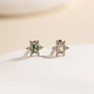 May include: A pair of silver-toned stud earrings featuring a star-shaped setting with pointed edges. Each earring contains a round, translucent stone with visible green moss-like inclusions, secured by a metallic prong setting.