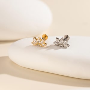 May include: Two stud earrings, one gold-toned and one silver-toned, each featuring a lotus flower design. The flower petals are adorned with clear, sparkling gemstones. The earrings are displayed on a white, rounded surface.