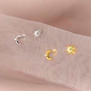 May include: A selection of celestial stud earrings. The earrings feature sun and crescent moon designs in silver and gold tones, some with small gemstones. The earrings are presented on a sheer, pale pink fabric.
