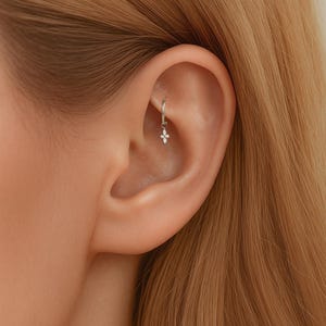 May include: A silver hoop earring with a small, dangling floral charm. The charm features four small, white gemstones arranged in a cross shape. The earring is worn in the helix piercing of an ear.