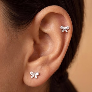 May include: A silver-toned butterfly earring stud featuring clear, faceted stones set in the wings, positioned on the earlobe, and a matching butterfly earring stud with clear stones located on the upper cartilage of the ear.
