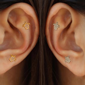 May include: Gold and silver-toned turtle stud earrings. The earrings are small, with a turtle design. The turtles are encrusted with small, clear stones. The earrings are shown on two ears.