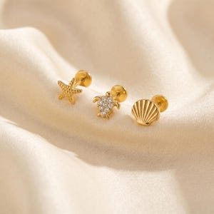 Può includere: Tre orecchini a lobo color oro con motivi marini. Un orecchino raffigura una stella marina testurizzata, uno una tartaruga marina con pietre pavé trasparenti sul guscio e uno una conchiglia con scanalature. Ogni pezzo ha una chiusura piatta.