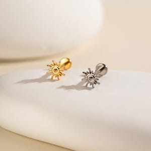 Peut inclure: Deux boucles d'oreilles en forme de soleil. L'une est dorée, l'autre argentée. Chaque boucle d'oreille a une tige et un petit dos rond. Le motif du soleil présente des pointes rayonnantes, créant un effet d'éclat. Les boucles d'oreilles sont présentées sur une surface blanche.
