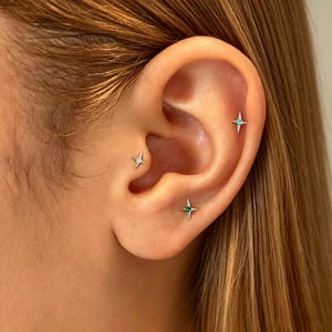 May include: Three silver star-shaped stud earrings are worn in the ear. The top earring features a light blue stone, the middle earring has a clear stone, and the bottom earring contains a small green stone, all set in a four-pointed star design.