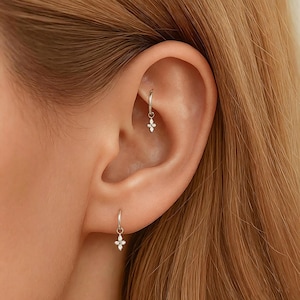 May include: Two silver hoop earrings, one in the lobe and one in the rook, each featuring a small, four-petaled flower charm. The charms are set with tiny, clear, sparkling stones that catch the light, creating a delicate and refined appearance.