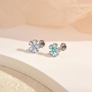 Può includere: Due orecchini a lobo a forma di fiore con chiusura piatta. Un orecchino presenta cinque pietre bianche iridescenti a goccia, mentre l'altro ha cinque pietre azzurre a goccia. Entrambi sono montati su una base in metallo color argento.