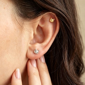 May include: A gold-toned cat-shaped stud earring positioned in the upper cartilage of the ear and a silver-toned cat-shaped stud earring located in the earlobe. Both earrings feature a detailed feline face design with pointed ears and whiskers.