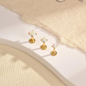 Può includere: Tre orecchini a lobo color oro con retro piatto di dimensioni variabili, ciascuno caratterizzato da una pietra rotonda bianca iridescente simile a un opale, fissata da quattro griffe. Presentano un perno sottile e una base circolare.